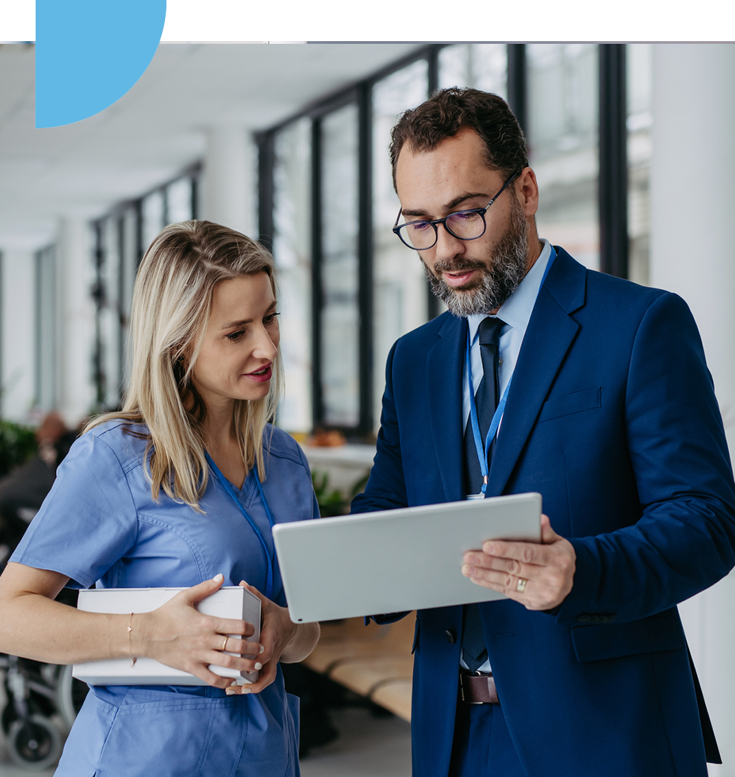 A female healthcare professional in scrubs and a male business professional in a blue suit are reviewing information on a digital tablet. They are standing in a modern, bright clinical or hospital hallway, engaged in a collaborative discussion.